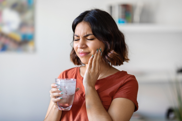 A woman holding a glass of ice water with her hand up to her face due to tooth sensitivity.
