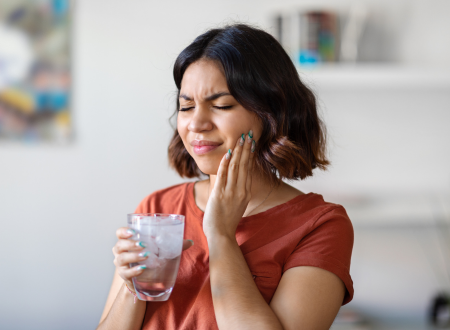 Woman holding a glass of water with her hand against her cheek due to tooth sensitivity and pain.